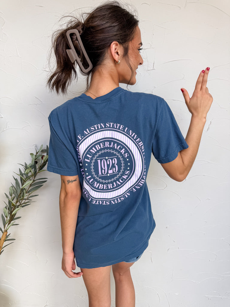 Person wearing a blue t-shirt with a circular design on the back, standing against a white wall.