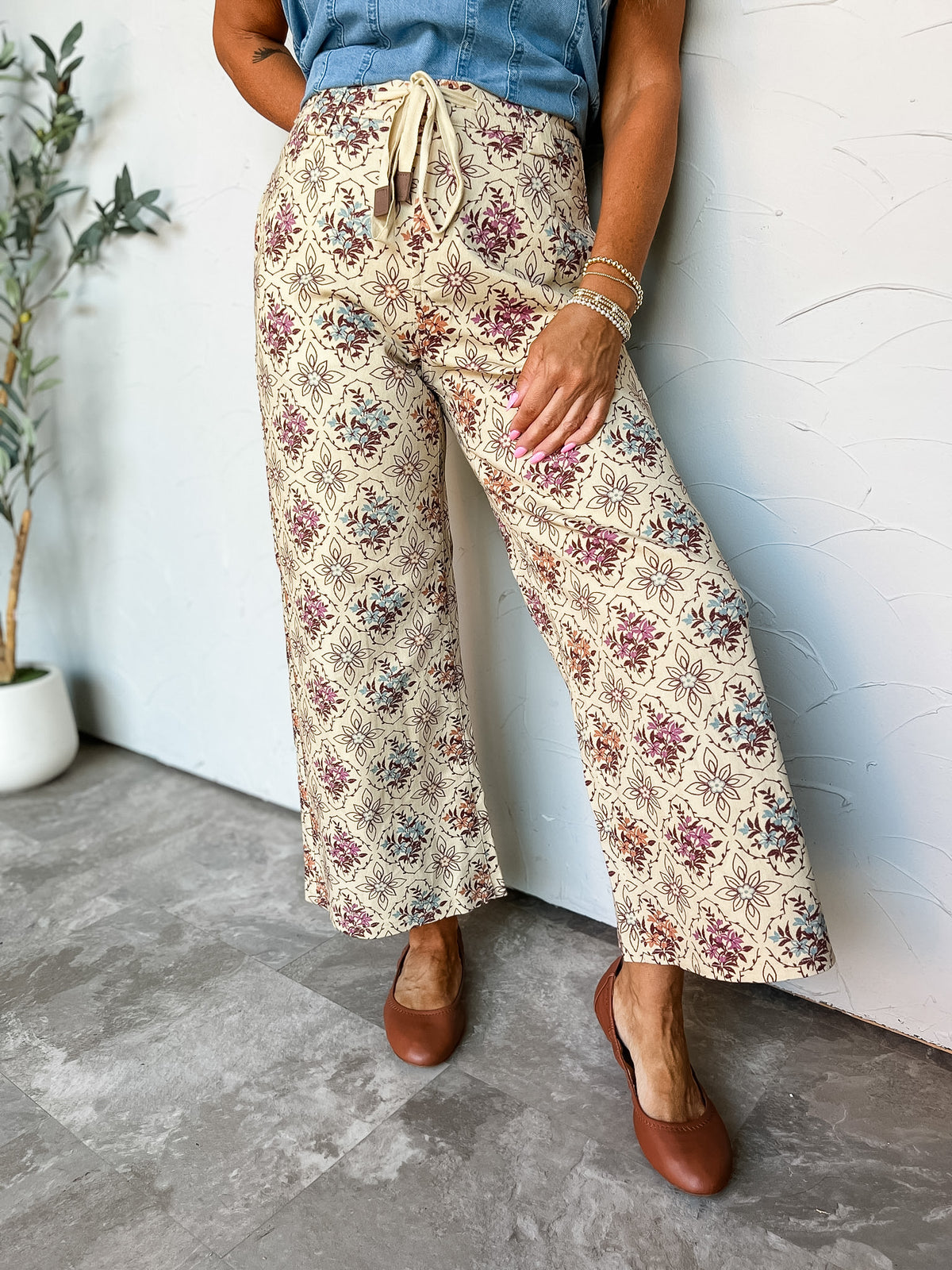 A waist-down photograph of a person leaning against a textured white wall wearing high-rise wide-leg pants made from a 97% cotton and 3% spandex blend, featuring an allover floral and geometric medallion pattern in burgundy, blue, and orange on a cream background, an adjustable cream drawstring waist tie, paired with a tucked-in medium-wash denim top, brown leather ballet flats, and beaded stretch bracelets.