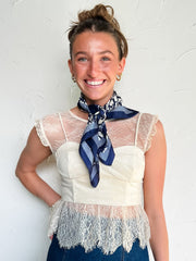 Woman wearing a white lace top and blue denim skirt with a blue scarf, standing against a white wall.