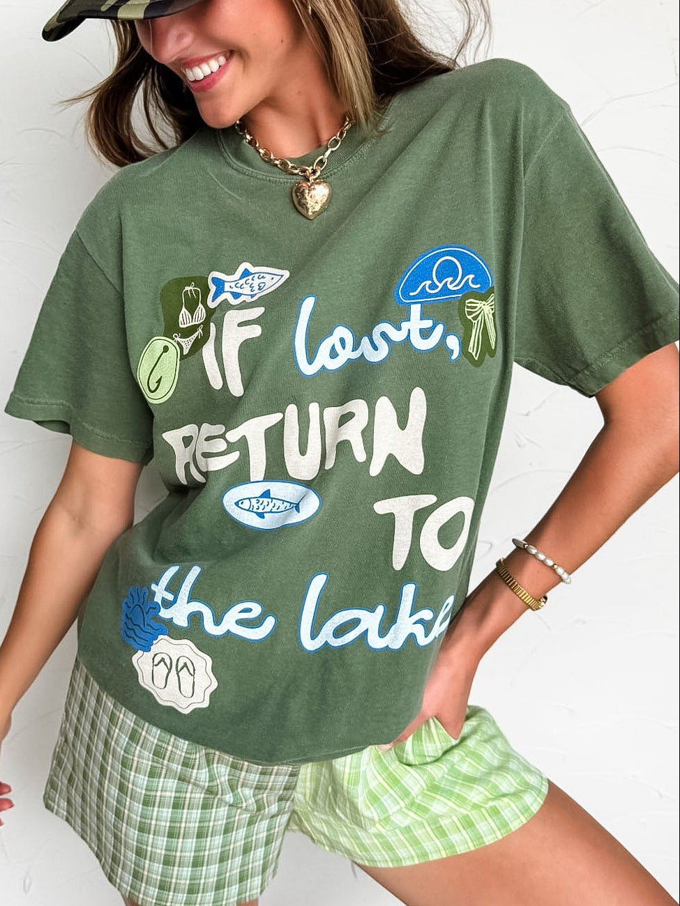 Woman wearing a green t-shirt with text and graphics, plaid shorts, and a camouflage cap.
