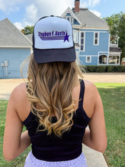 A woman viewed from behind wearing a personalized white and navy trucker hat with the text 'Stephen F. Austin' and a purple star, set against a background of a blue house and green lawn.