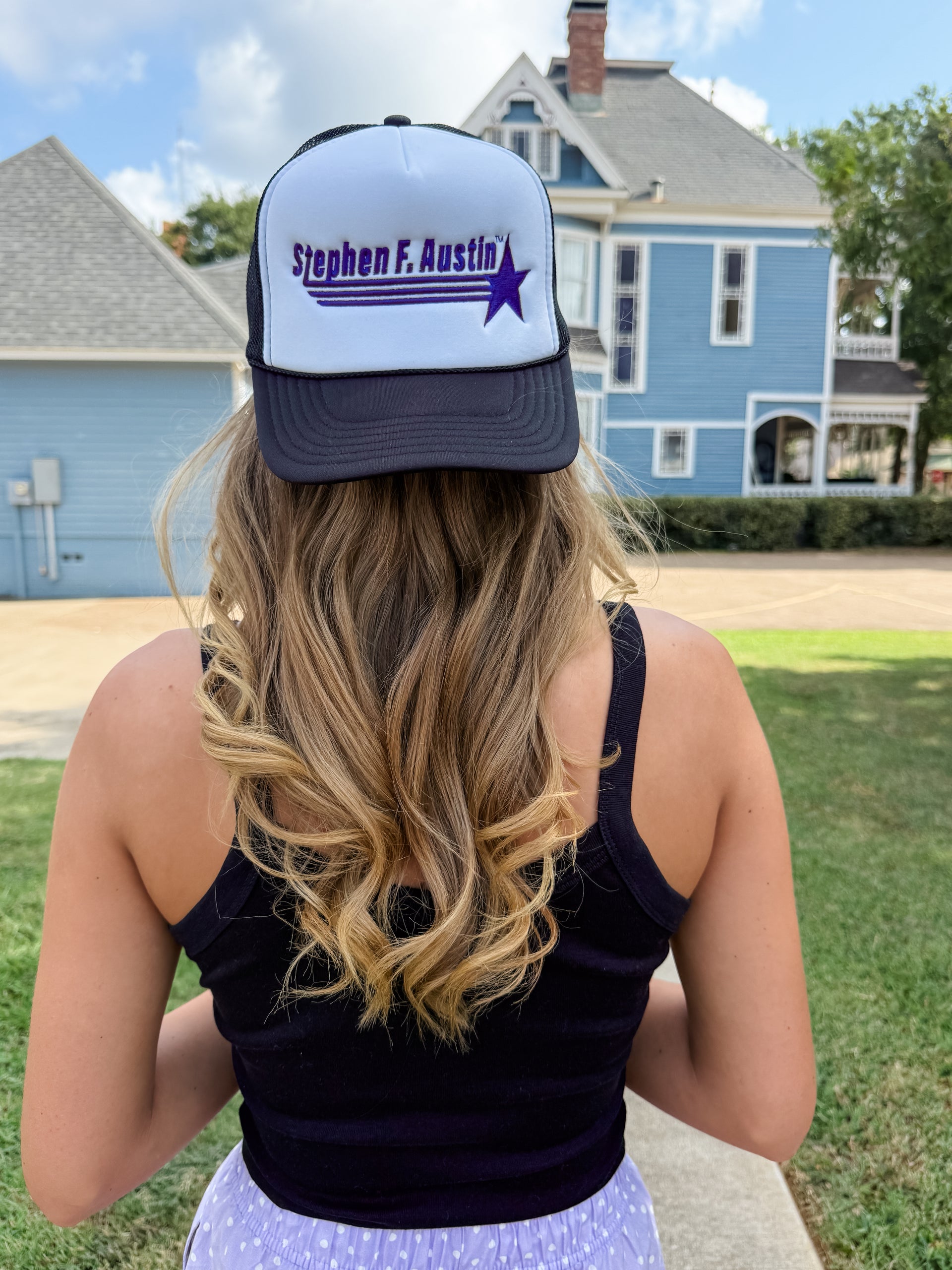 A woman viewed from behind wearing a personalized white and navy trucker hat with the text 'Stephen F. Austin' and a purple star, set against a background of a blue house and green lawn.