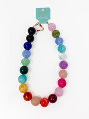 A multicolor beaded necklace with large round acrylic beads in a rainbow gradient, displayed on a white background with a turquoise brand tag attached.