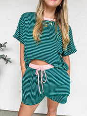 A three-quarter length photo of a smiling blonde woman leaning against a textured white wall. She is wearing a green and teal fine-striped matching lounge set that includes a short-sleeved top with a contrasting light pink crew neckline. The top is loose, subtly revealing the light pink elastic waistband and matching drawstring tie of the coordinating high-waisted shorts. She has one hand in her pocket and is wearing a gold chain necklace with charms.