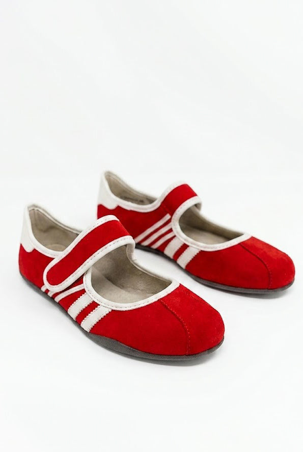 A pair of red suede Mary Jane style flats with white trim and white athletic stripes on the sides, set against a solid white background. The shoes feature an adjustable hook-and-loop Mary Jane strap across the top and a thin, low-profile black sole.
