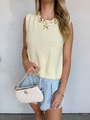A woman wearing a buttercream yellow sleeveless vest made of a 100% acrylic knit construction, featuring a round crew neckline, a finely textured body, and a ribbed hemline. The top showcases a dramatic open-back design worn over a tan tube top, complete with a self-tie closure at the nape of the neck with long dangling knit straps framed by vertical ribbing. She is styled against a textured off-white wall wearing light-wash denim shorts, a chunky gold chain and pearl necklace with a large starfish charm, a