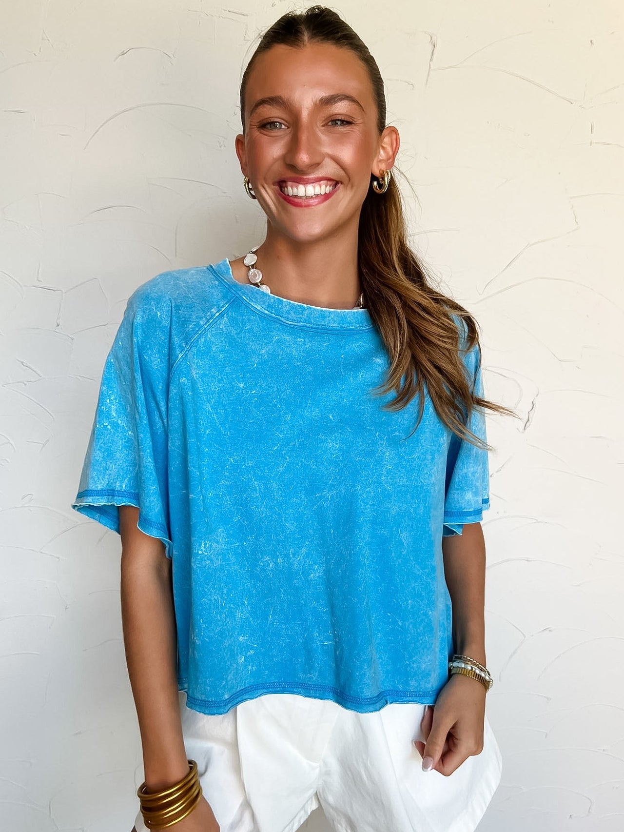 Woman wearing a blue acid washed cropped shirt and white shorts against a light background