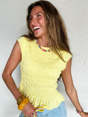 Woman wearing a yellow top with colorful necklace beads and denim shorts against a white textured wall.