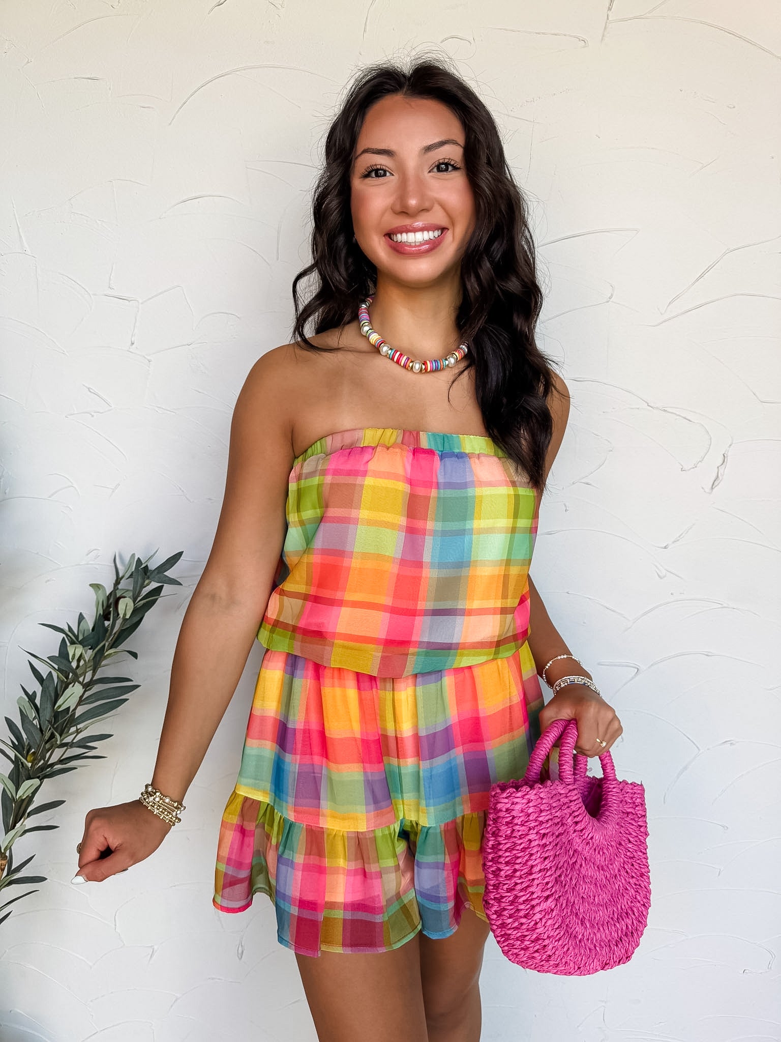 A close-up view of a person standing against a textured white wall, wearing a strapless multi-color plaid romper made from 100% polyester. The romper features a vibrant grid pattern in shades of pink, orange, yellow, green, blue, and purple, complete with an elasticized waist and a tiered ruffle hem. She is accessorized with a colorful beaded collar necklace, stacked beaded bracelets on both wrists, and a bright pink woven straw tote bag.