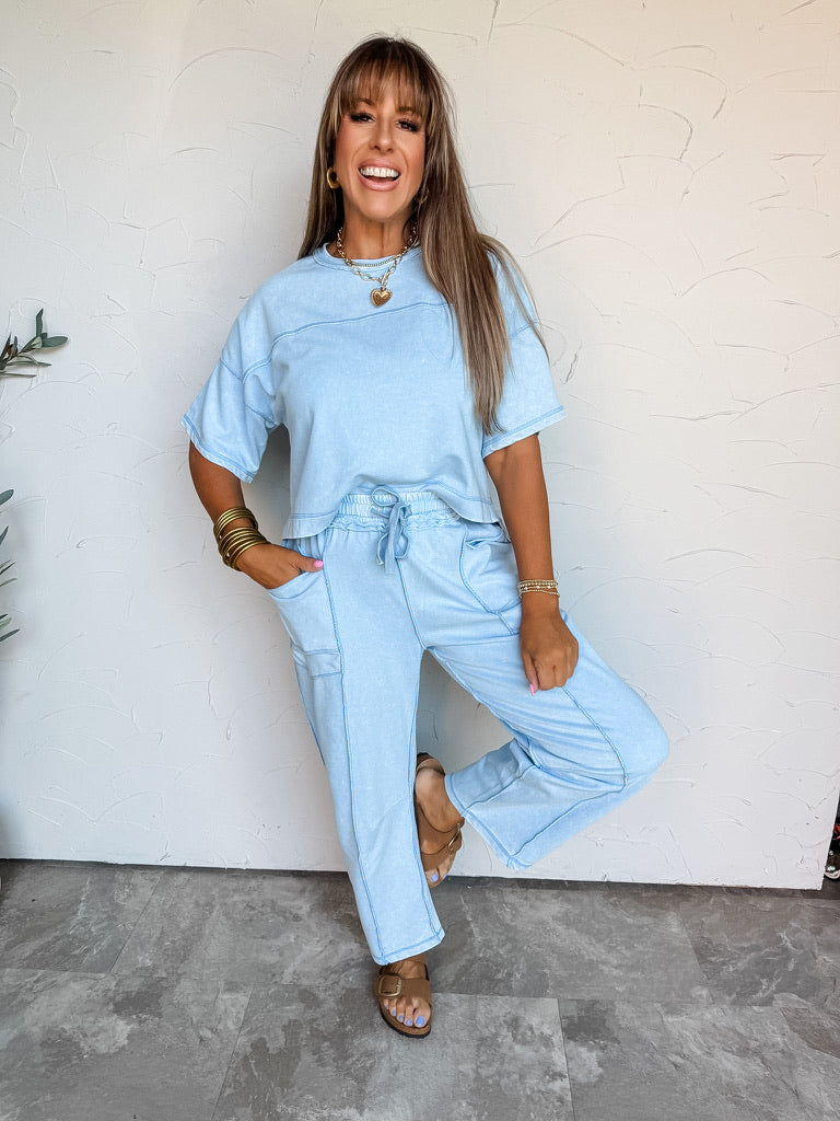 A full-length photograph of a smiling woman with straight light-brown hair and front bangs holding a strand of her hair and posing on one leg against a textured white wall. She is wearing a matching two-piece light blue lounge set featuring a relaxed short-sleeved crewneck top with exposed seam detailing, paired with matching straight-leg pants with an elastic drawstring waist and side pockets, styled with brown slide sandals, stacked gold bangles, gold hoop earrings, and a gold chain heart pendant necklace