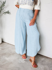 A photograph focusing from the waist down of a person leaning against a textured white wall. They are wearing light-wash, flowy wide-leg pants made from 100% Lyocell fabric, featuring raised vertical front center seams and subtly rounded side hem detailing, styled with flat metallic strap sandals and a cream graphic top.