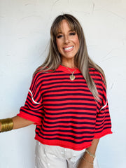A waist-up photograph of a smiling woman with straight light-brown hair and front bangs posing against a textured white wall. She is wearing a short-sleeved fine knit sweater made from a 50% viscose, 30% polyester, and 20% nylon blend, featuring horizontal red and navy blue stripes, white piping across the drop-shoulder seam, wide elbow-length sleeves, and a ribbed crew neck, paired with cream polka dot pants and styled with stacked gold bangles and a layered gold heart pendant necklace.