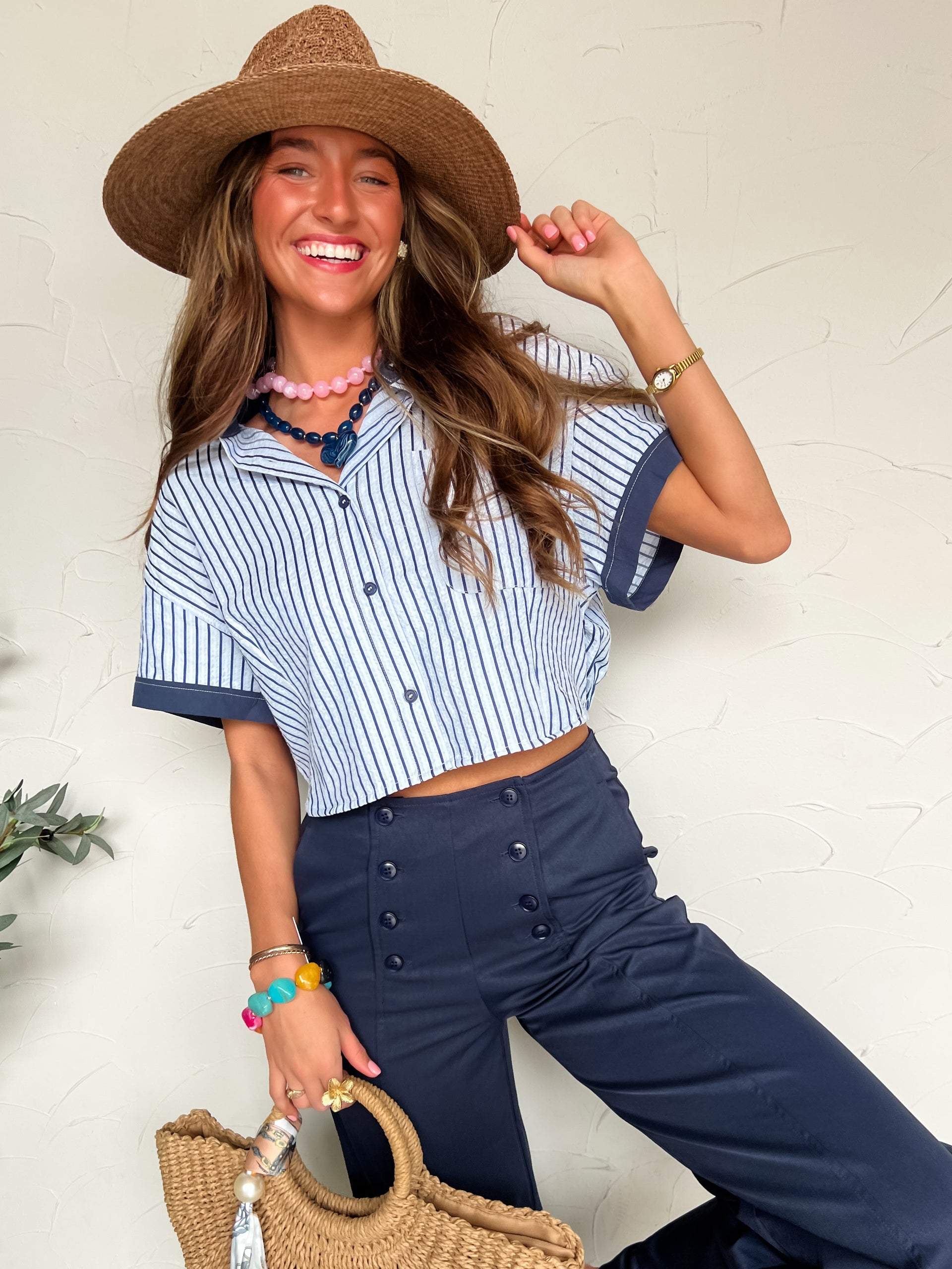 Woman wearing a striped shirt and wide-leg pants with a straw hat and bag, standing against a light background.