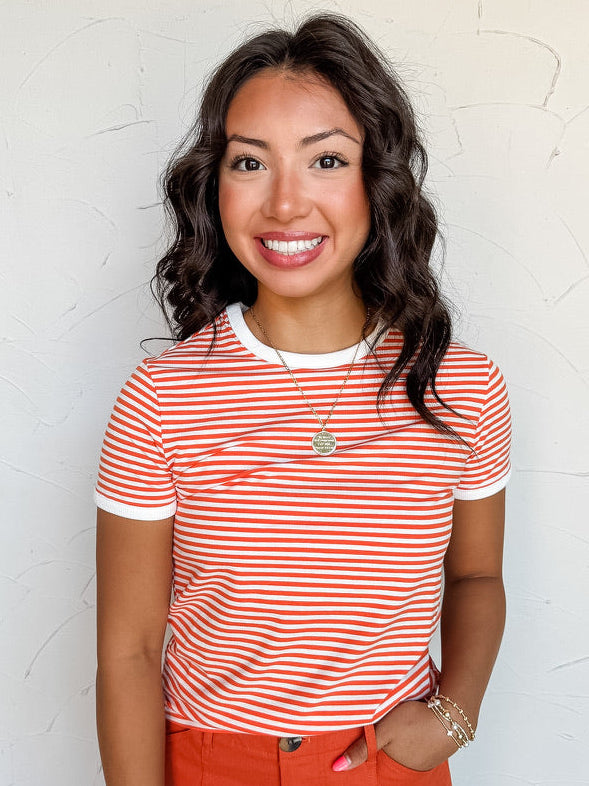 A three-quarter length photograph of a smiling woman with dark wavy hair posing with one hand in her pocket against a textured white wall. She is wearing a fitted orange and white horizontal micro-striped short-sleeved ringer t-shirt made from a 95% cotton and 5% spandex blend, featuring white contrast trim along the round neckline and sleeve cuffs, tucked into bright orange wide-leg pants and styled with a gold pendant necklace and delicate beaded bracelets.