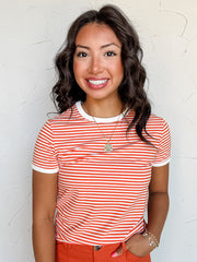 A three-quarter length photograph of a smiling woman with dark wavy hair posing with one hand in her pocket against a textured white wall. She is wearing a fitted orange and white horizontal micro-striped short-sleeved ringer t-shirt made from a 95% cotton and 5% spandex blend, featuring white contrast trim along the round neckline and sleeve cuffs, tucked into bright orange wide-leg pants and styled with a gold pendant necklace and delicate beaded bracelets.