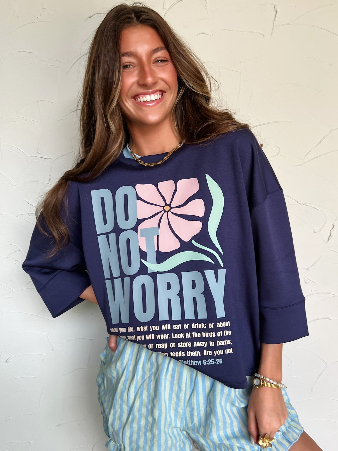 Woman wearing a navy sweatshirt with 'Do Not Worry' text and floral design, standing against a textured white wall.