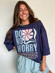Woman wearing a navy sweatshirt with 'Do Not Worry' text and floral design, standing against a textured white wall.