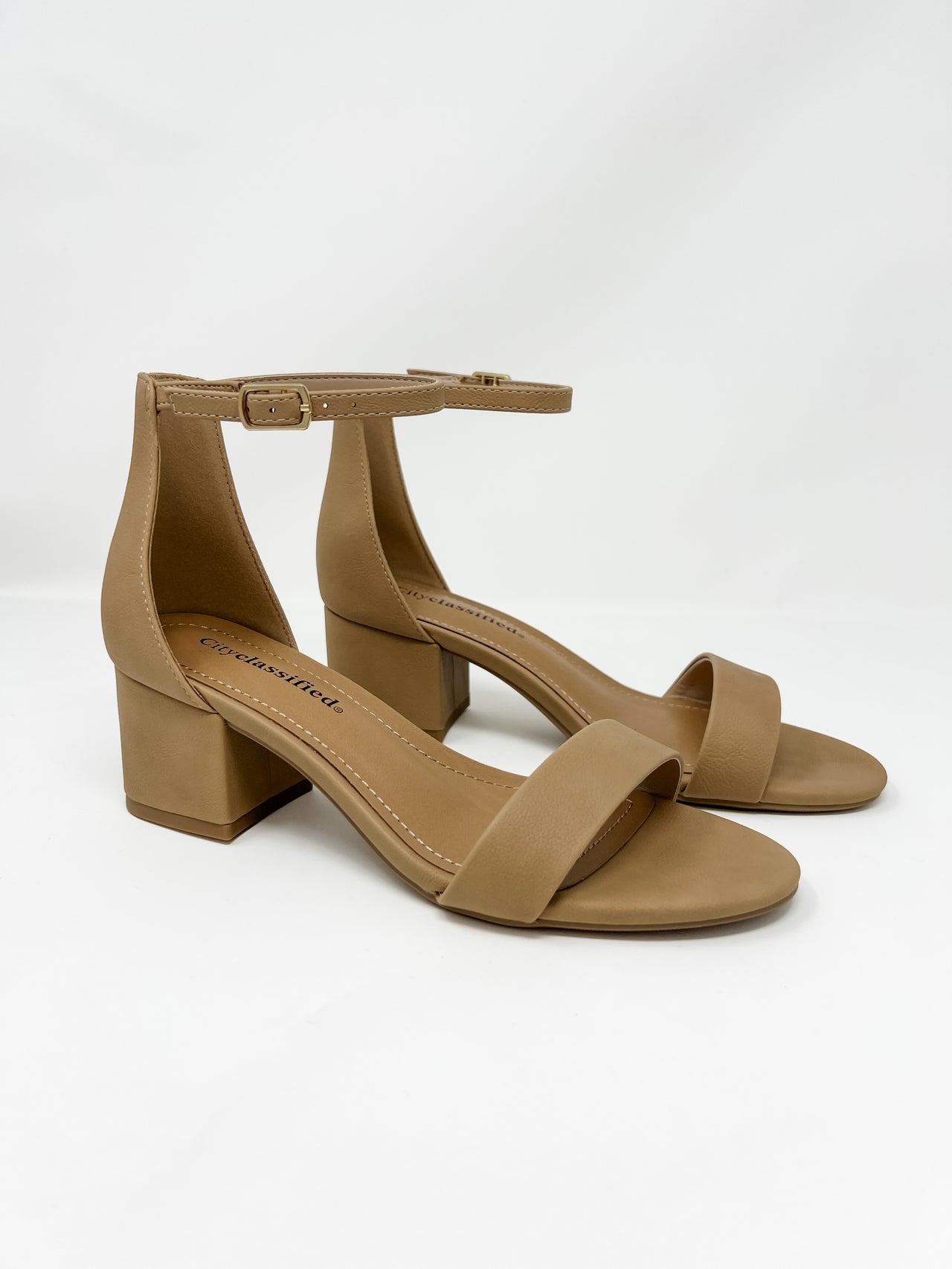 A pair of City Classified brand block heel sandals in a warm nude tan color, shown against a solid white background. The shoes feature a single toe strap, an adjustable ankle strap with a small gold buckle, a high closed back, and a low, chunky block heel.