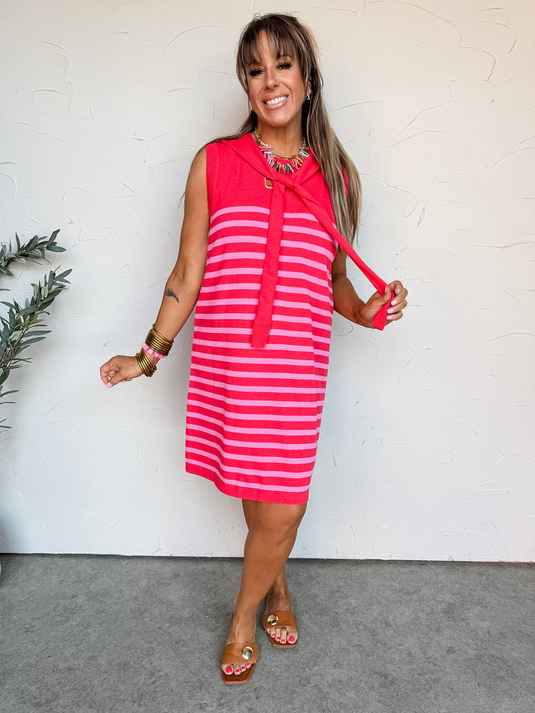 A full-length front view and a back view of a smiling woman with brown hair leaning against a textured off-white wall, wearing a sleeveless knit shift dress. The mini-length dress is crafted from a soft, fine-knit 63% viscose and 37% nylon blend fabric featuring an all-over vibrant pink and red horizontal stripe pattern. It is uniquely detailed with an attached solid red knit collar that mimics a sweater draped over the shoulders, falling in a square flap across the back 