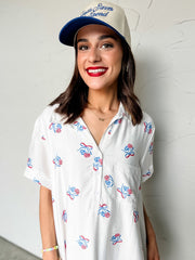 Woman wearing a white dress with colorful patriotic patterns and a blue cap against a light gray wall.
