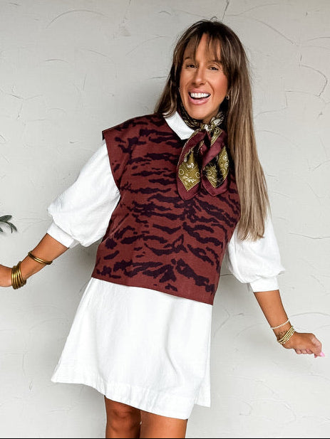 A full-length photograph of a smiling woman with long brown hair with bangs holding her arms slightly outward while posing against a textured white wall next to a potted olive tree and woven basket. She is wearing a white 100% polyester t-shirt mini dress with a pointed collar, a buttoned front placket, a chest patch pocket, dropped shoulders, and elbow-length puff sleeves, styled with tan suede ankle boots, a chunky pearl necklace featuring a gold heart pendant, stacked gold bangles, and delicate gold chai