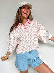 Woman wearing a striped pink, navy and white shirt and denim shorts against a white wall.