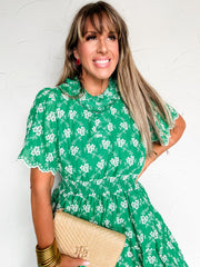 Woman wearing a green floral dress holding a beige clutch against a white background