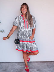 A woman modeling a short-sleeve tiered dress with a black and white floral pattern and red trim, standing against a white wall.