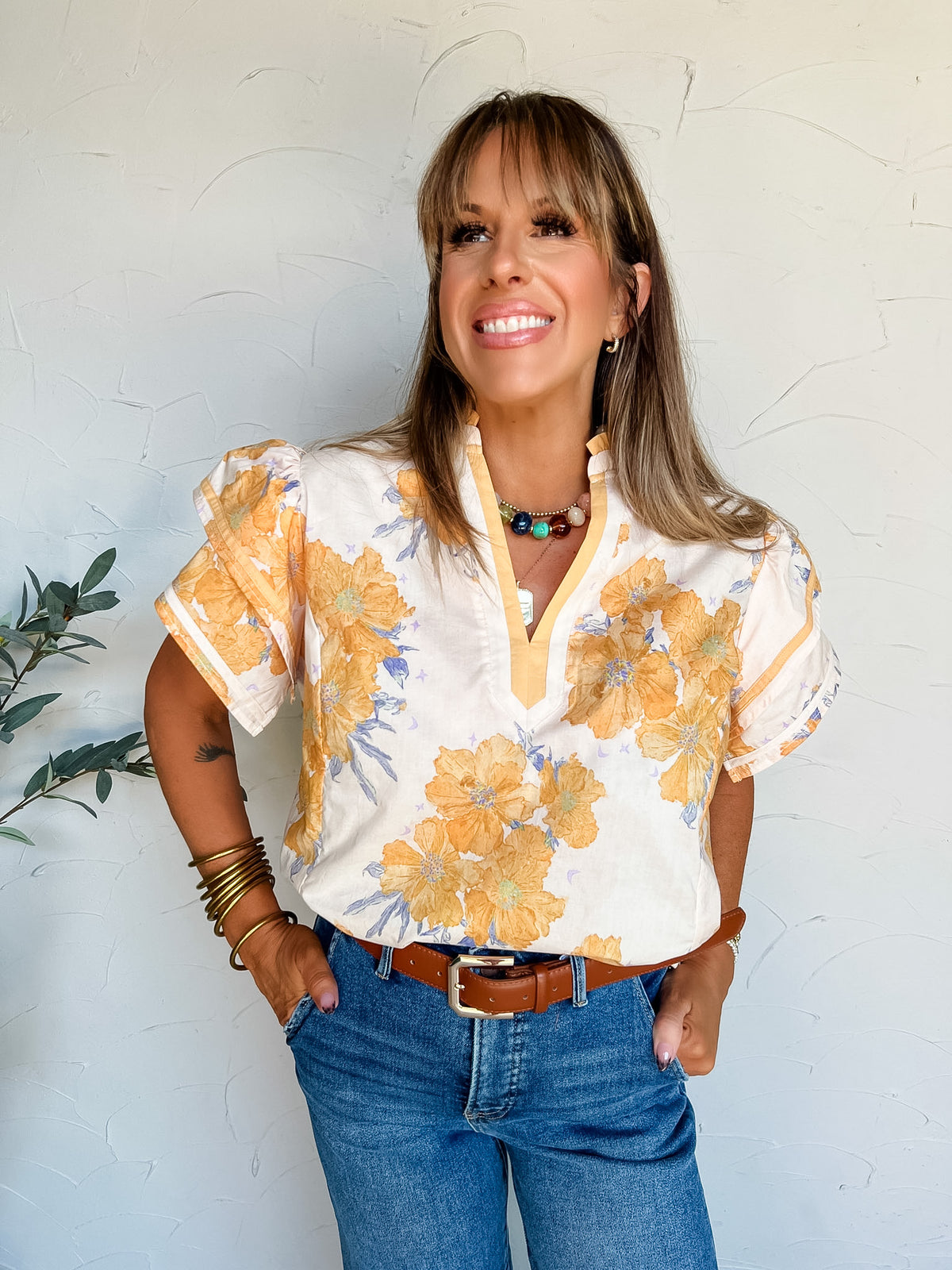A portrait photograph of a smiling woman with straight blonde hair and bangs looking off to the side with her hands in her pockets against a textured off-white wall next to a potted olive plant. She is wearing a short-sleeved 100% cotton blouse with a yellow floral and purple leaf print, featuring a split neck with yellow trim, a mandarin collar, and banded flutter sleeves, tucked into medium-wash blue jeans with a brown leather belt, styled with a colorful stone beaded choker necklace, a gold pendant, stac