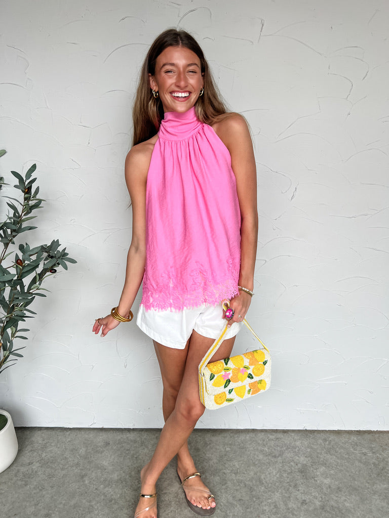 Woman wearing a pink top and white shorts, holding a yellow clutch with floral design, against a white wall.
