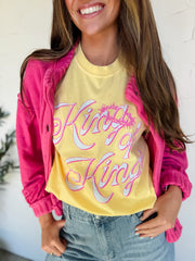 Person wearing a yellow 'King of Kings' shirt with a pink jacket and jeans indoors.