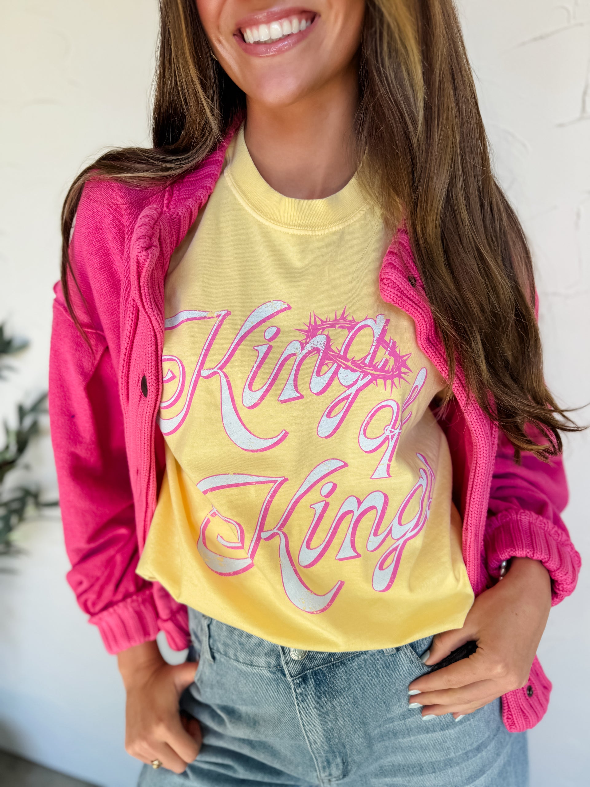 Person wearing a yellow 'King of Kings' shirt with a pink jacket and jeans indoors.