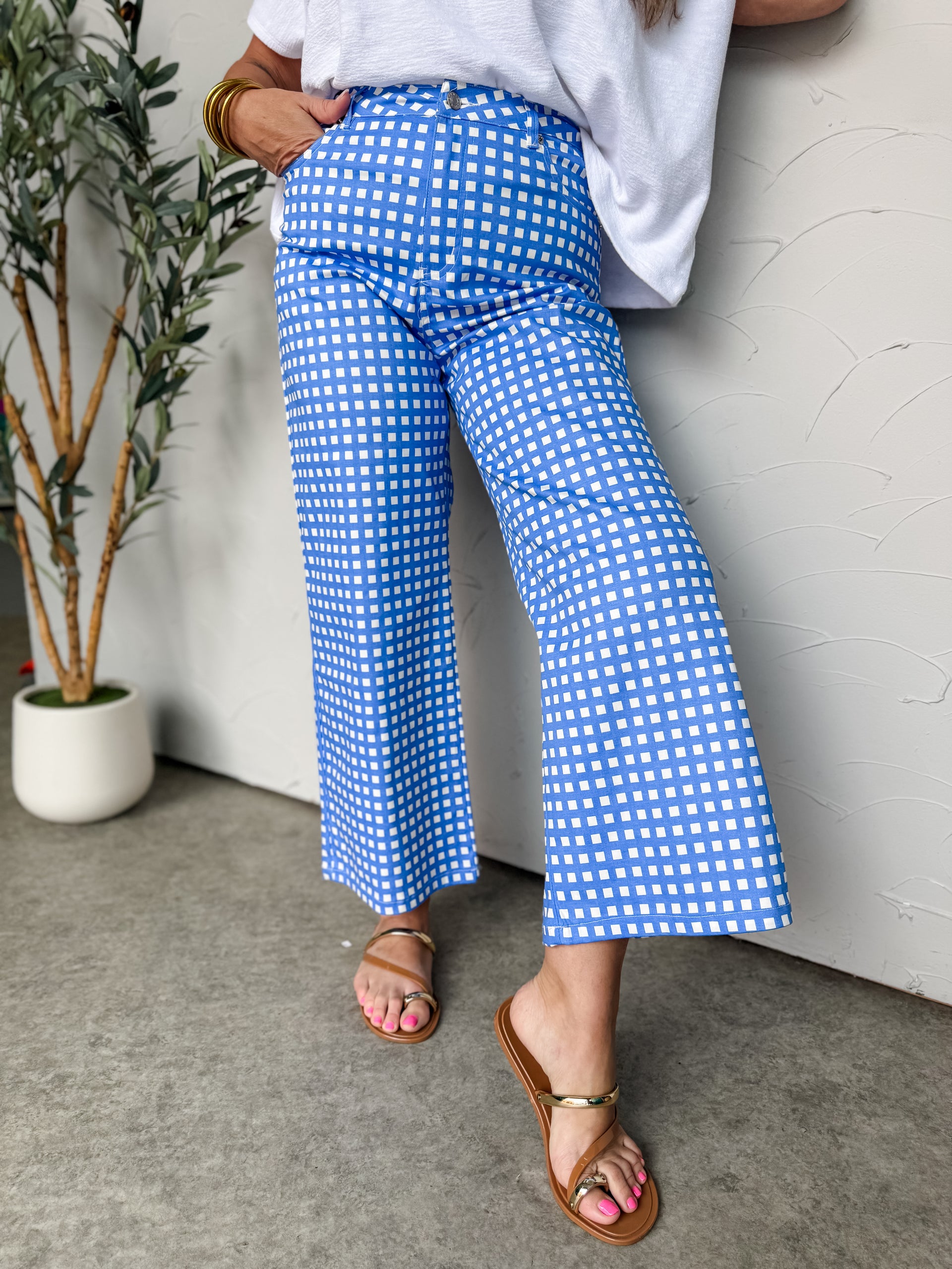 A pair of high-rise, wide-leg gingham pants in a vibrant blue and white checkered pattern, crafted from 100% cotton. The pants feature an ankle-length cropped fit, a structured waistband with belt loops, a button closure with a zip fly, and classic five-pocket detailing.