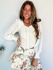 Woman wearing a white blouse with long sleeves and a floral skirt against a light blue wall.