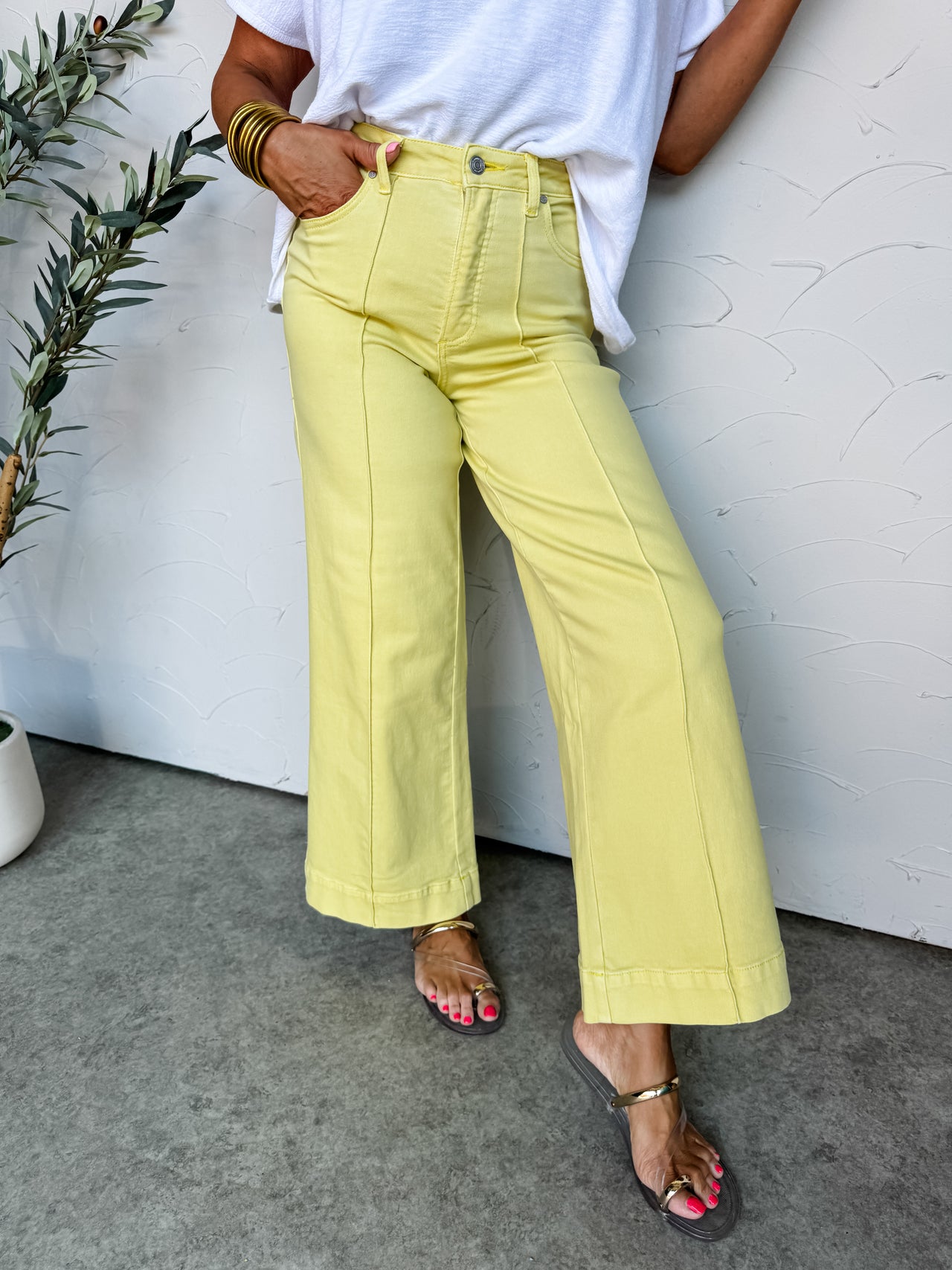 A lower-body view of a person standing against a textured off-white wall, wearing pale yellow wide-leg pants. The high-waisted trousers are crafted from a stretch denim blend of 91% cotton, 6% polyester, and 3% spandex, featuring a silver button closure, a zip fly, standard pockets, and prominent vertical seams down the center of each leg. They are styled with a tucked-in white textured top and metallic gold flat sandals.