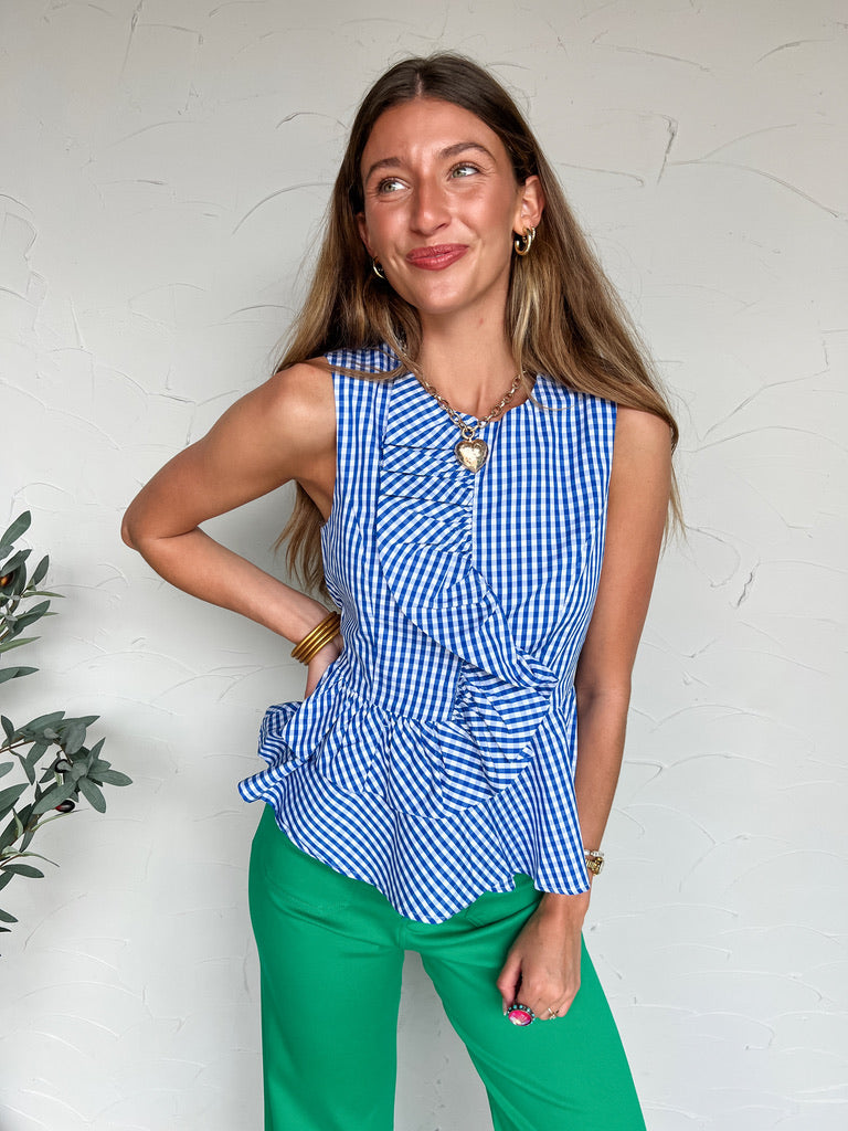 Woman wearing a blue and white checkered top and green pants against a white wall.