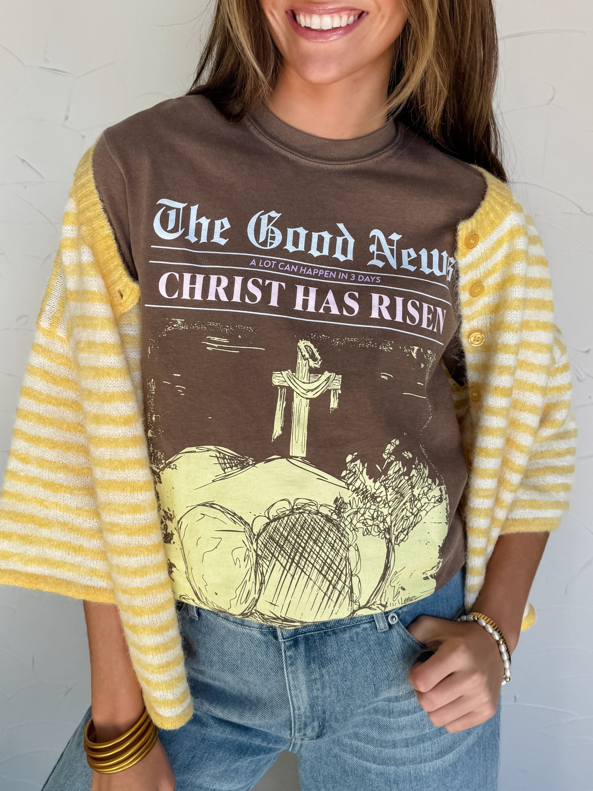 Woman wearing a graphic t-shirt with text "Christ has risen" newspaper graphic and design, standing against a white wall.