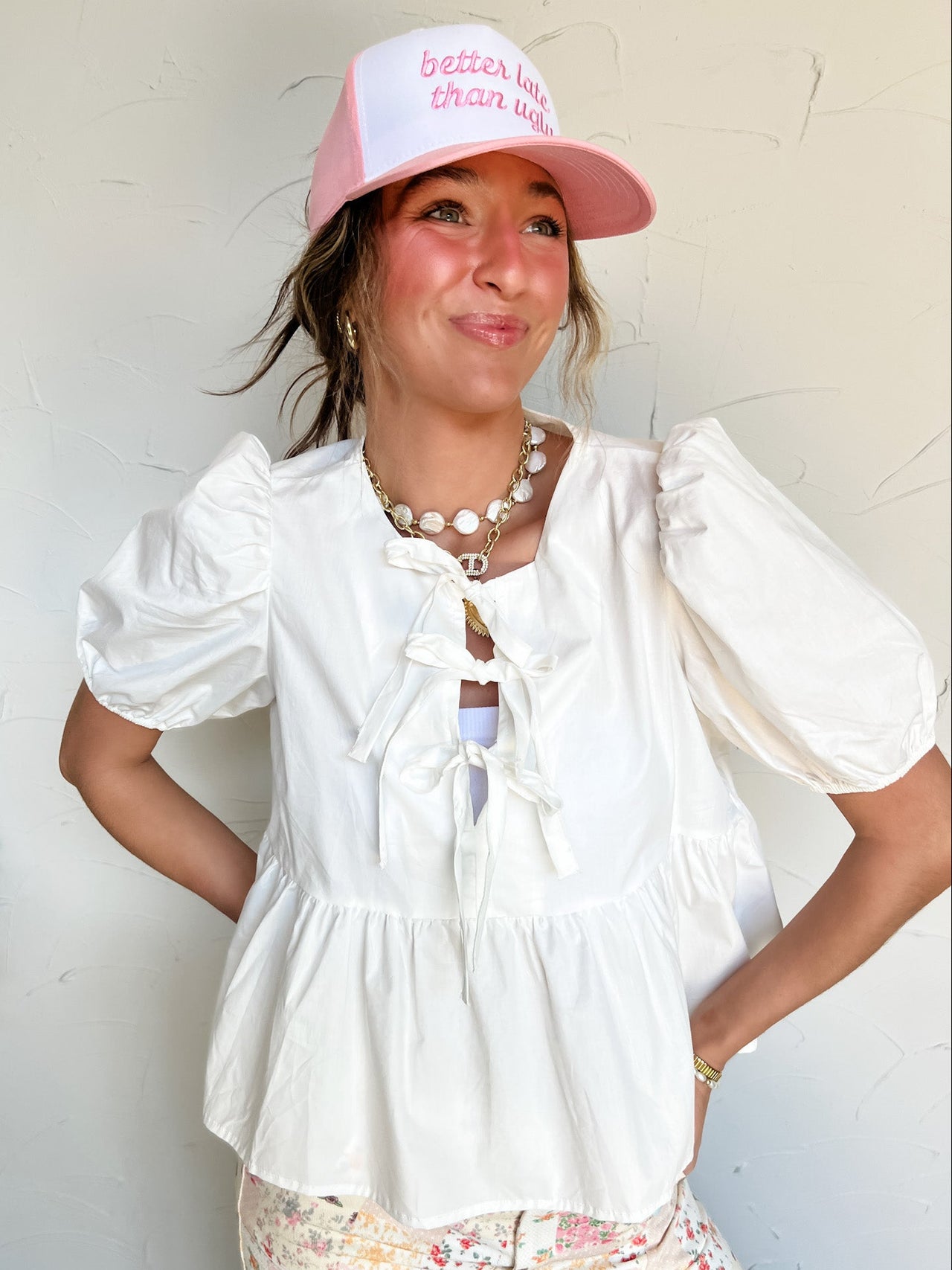 Woman wearing a white peplum top with bows and pink cap with text against a light background