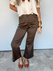 Model wearing Be On My Way Brown Pants by Umgee, made from 65% rayon, 30% nylon, and 5% spandex, featuring a rich brown color, front button closure, belt loops, front pockets, and cropped, slightly flared legs, styled with a cream ruffled blouse and brown shoes.


