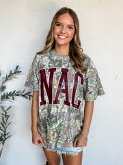 A woman wearing a camouflage graphic t-shirt with 'NAC Texas' printed in maroon and white, paired with light blue denim skirt, standing against a white textured wall.