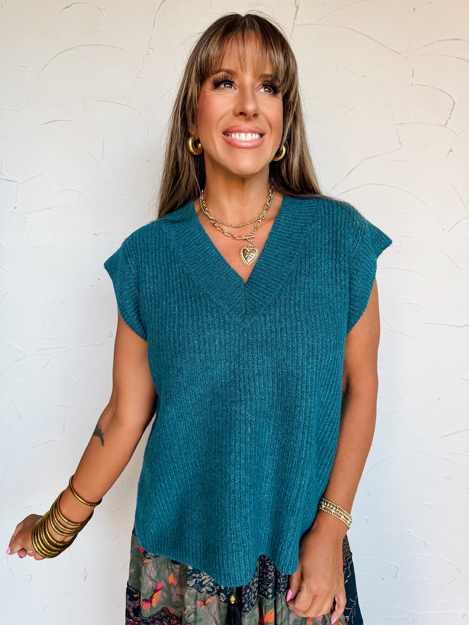 A front-facing waist-up shot of a smiling woman with straight light-brown hair and front bangs posing against a textured white wall. She is wearing a teal blue knit sweater vest made from a 98% polyester and 2% spandex blend, featuring cap-sleeve shoulders, a deep ribbed V-neckline, and fine vertical ribbed texture, accessorized with gold hoop earrings, a layered gold chain necklace with a heart pendant, and stacked gold bangles.