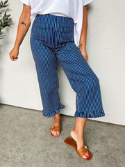 A lower-body shot of a person standing against a textured off-white wall, wearing high-waisted pinstripe trousers crafted from a 98% cotton and 2% spandex blend fabric. The navy blue pants feature fine vertical white stripes, a smooth front waistband, a straight-leg silhouette, and playful ruffled trim at the cropped ankle-length hems. The pants are paired with a textured white short-sleeve top tucked into the waistband and styled with brown flat slide sandals adorned with large gold statement buttons