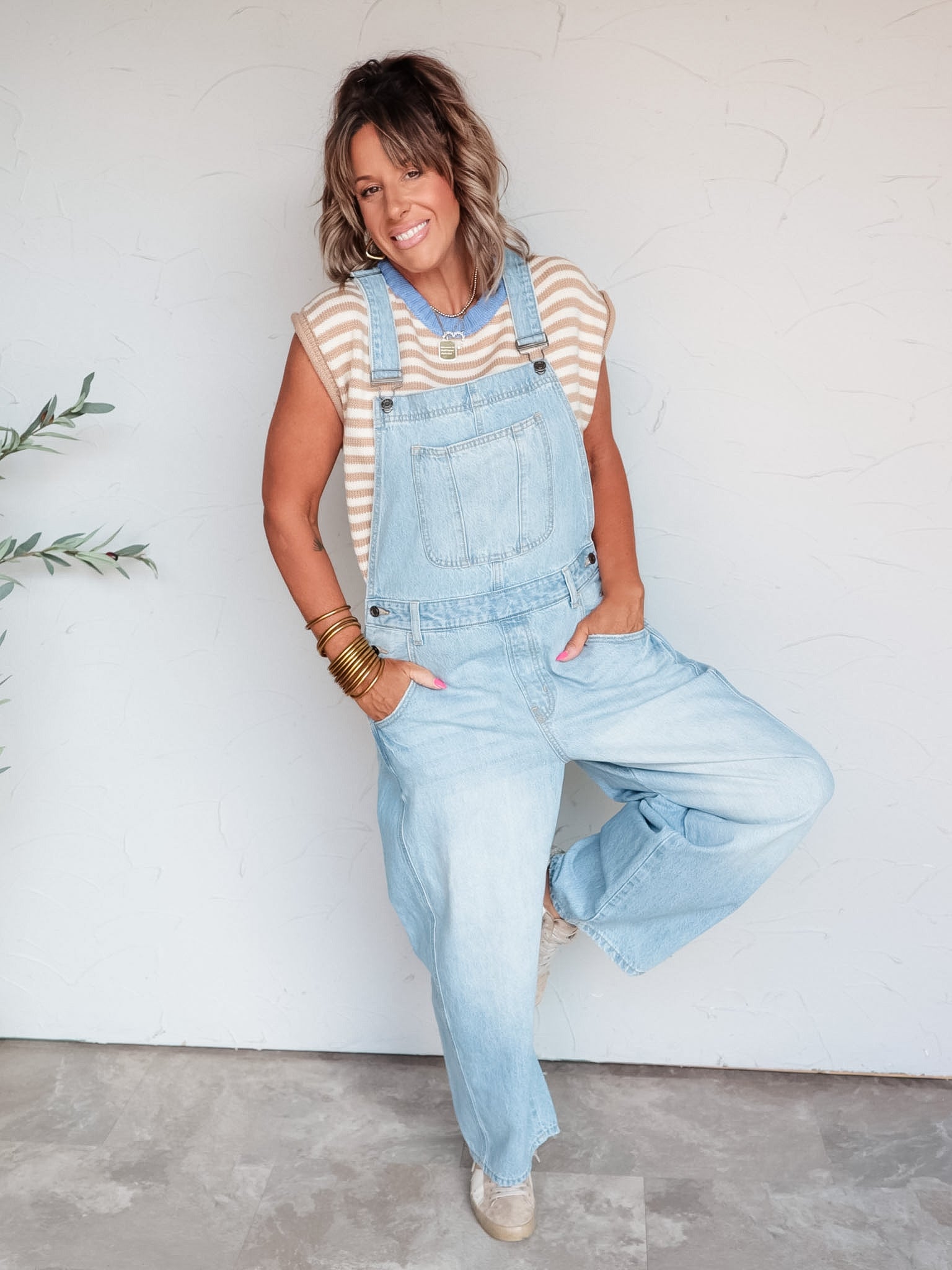 A full-length photograph of a smiling woman with medium-length brown hair and bangs posing with her hands in her pockets and one leg raised against a textured white wall. She is wearing light-wash denim overalls made from 100% cotton fabric, layered over a tan and white striped knit sweater vest with a light blue neck trim, paired with low-top sneakers and styled with layered pendant necklaces, gold hoop earrings, and stacked gold bangles.