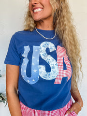 A person wearing a navy blue t-shirt with a graphic that reads 'USA' in white and red letters, and red plaid boxer shorts, paired with a white cap.