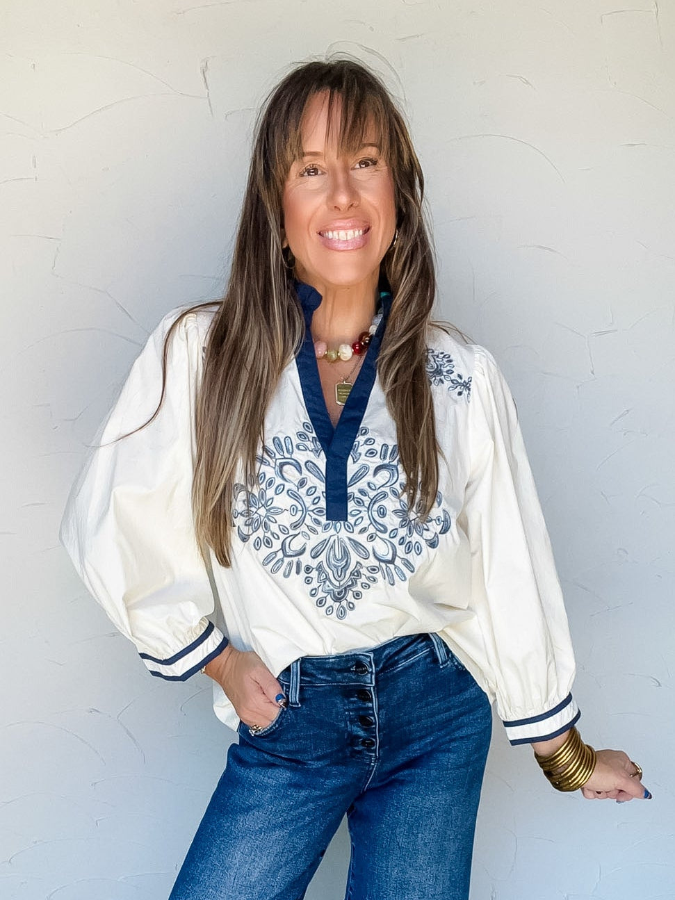 Model wearing the Hold It Together Floral Smocked Ruffle Top by Umgee in 100% polyester, featuring navy floral and paisley motifs on a textured ivory background, a ruffled neckline with a V-shaped opening, smocked detailing, and short puff sleeves with gathered cuffs, styled with dark blue button-front jeans and gold-tone jewelry
