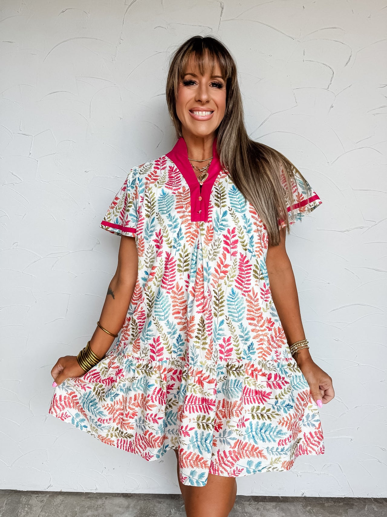 A front-facing photograph of a smiling woman with straight light-brown hair and front bangs holding the sides of her dress hem against a textured white wall. She is wearing an A-line mini dress made from 100% cotton, featuring a colorful leaf print in shades of pink, teal, orange, and green on a white base, a vibrant magenta split-neck stand collar with two buttons, short flutter sleeves with magenta piping, and a gathered ruffle hem, accessorized with stacked gold bangles and a gold chain necklace.