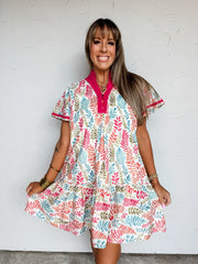 A front-facing photograph of a smiling woman with straight light-brown hair and front bangs holding the sides of her dress hem against a textured white wall. She is wearing an A-line mini dress made from 100% cotton, featuring a colorful leaf print in shades of pink, teal, orange, and green on a white base, a vibrant magenta split-neck stand collar with two buttons, short flutter sleeves with magenta piping, and a gathered ruffle hem, accessorized with stacked gold bangles and a gold chain necklace.