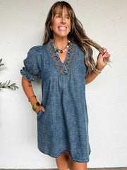 A three-quarter length photograph of a smiling woman with long brown hair and bangs holding a strand of her hair while posing with one hand in her pocket against a textured off-white wall. She is wearing a short-sleeved acid-wash denim shift dress made from a 90% cotton and 10% polyester blend, featuring a split V-neckline detailed with mustard yellow zigzag embroidery, short puff sleeves with smocked ruffle cuffs, and side pockets, styled with a colorful beaded necklace, stacked brass bangles, and beaded s
