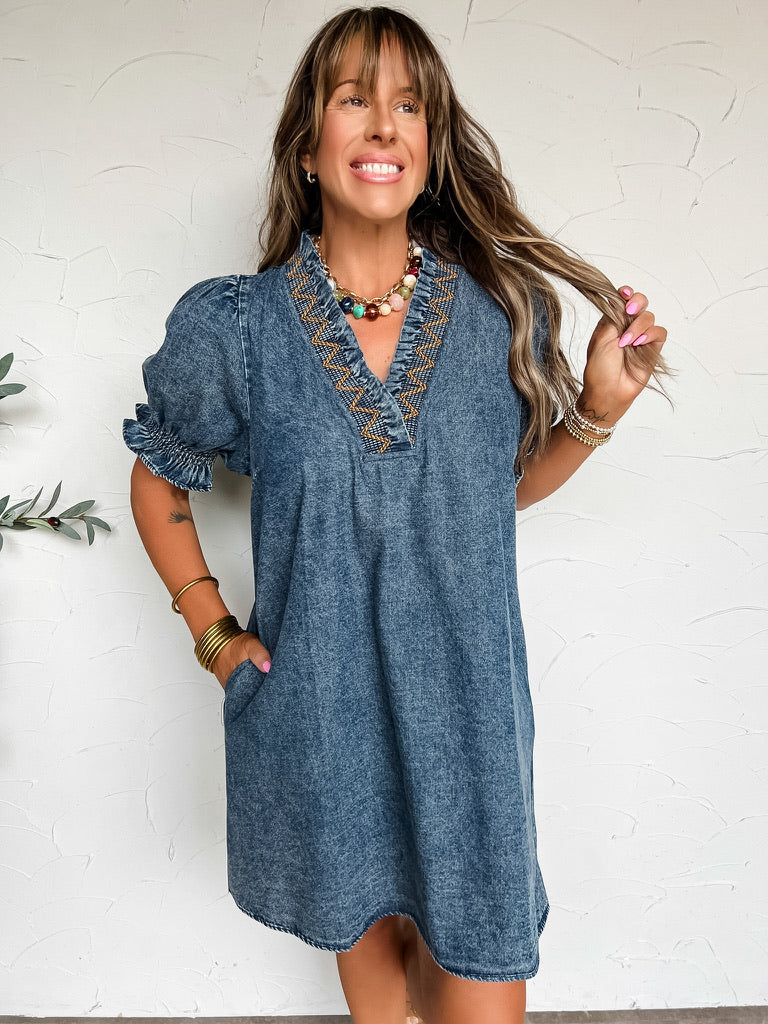 A three-quarter length photograph of a smiling woman with long brown hair and bangs holding a strand of her hair while posing with one hand in her pocket against a textured off-white wall. She is wearing a short-sleeved acid-wash denim shift dress made from a 90% cotton and 10% polyester blend, featuring a split V-neckline detailed with mustard yellow zigzag embroidery, short puff sleeves with smocked ruffle cuffs, and side pockets, styled with a colorful beaded necklace, stacked brass bangles, and beaded s