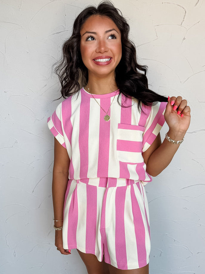 A three-quarter length photograph of a smiling woman with dark wavy hair touching a strand of her hair while posing against a textured white wall. She is wearing a short-sleeved top made from a 58% polyester, 30% cotton, 10% rayon, and 2% spandex blend, featuring wide pink and off-white vertical stripes, a chest pocket, and cuffed cap sleeves, paired with matching striped shorts and styled with a round pendant necklace and gold beaded bracelets.